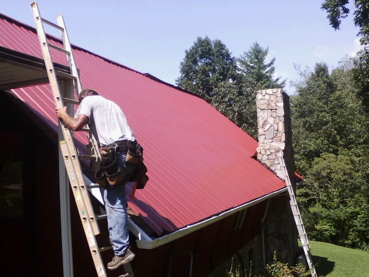 Roofer on a ladder beside a red metal roof and stone chimney — Soffit & Fascia Repair by Roofing Brothers in Douglasville